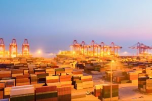 Containers lined up in a part at dawn, with lights over them.